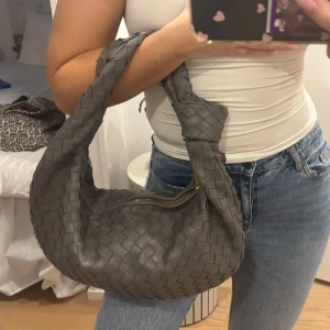 Grå flätad axelväska - Snygg grå axelväska med flätat mönster och dragkedja. Perfekt för att ge din outfit en stilren touch. Väskan har en knuten detalj på axelremmen som ger den en unik look.