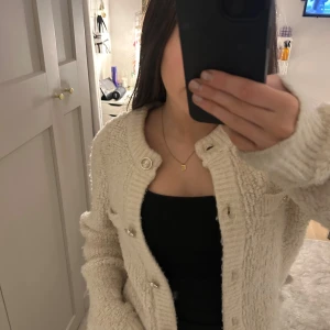 Beige stickad kofta med knappar - Mysig beige stickad kofta med dekorativa knappar framtill. Koftan har en rund halsringning och långa ärmar, perfekt för kyligare dagar. Den har en klassisk och stilren design som passar till många olika outfits.