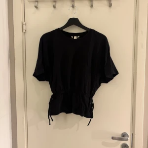 Svart kortärmad topp med knyt - Säljer en svart kortärmad topp med knyt i midjan. Perfekt för en stilren look. Toppen har en rund hals och är i ett mjukt material.