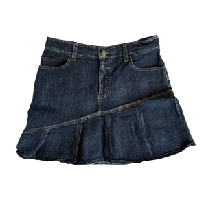 Mörkblå jeanskjol - Snygg mörkblå jeanskjol med en unik snedskuren design och volangdetalj. Kjolen har klassiska femfickor och en knappstängning fram. Perfekt för en avslappnad stil.