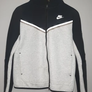 Grå och svart jacka från Nike - Säljer en snygg grå och svart jacka från Nike med dragkedja och hög krage. Jackan har ett stilrent mönster med vita och svarta detaljer samt Nike-logga på bröstet. Perfekt för en sportig look!