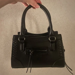 Svart handväska med dragkedja från Gina  - Snygg svart handväska i skinn med stilrena detaljer. Väskan har en praktisk dragkedja framtill och två robusta handtag. Perfekt för att bära dina nödvändigheter med stil. En klassisk accessoar som passar till många tillfällen. Pris kan diskuteras! 