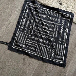 Svart halsduk från Emporio Armani - Snygg svart halsduk från Emporio Armani med ett stilrent mönster av vita logotyper. Perfekt accessoar för att lyfta din outfit. Tillverkad i ett mjukt och bekvämt material. Använd max 3 ggr