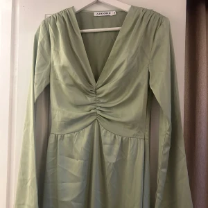 Grön långklänning från Adoore - Elegant grön långklänning från Adoore med v-ringning och rynkdetaljer framtill. Klänningen har långa ärmar och är tillverkad i ett mjukt, glansigt material som ger en lyxig känsla. Perfekt för speciella tillfällen. Aldrig använd. 