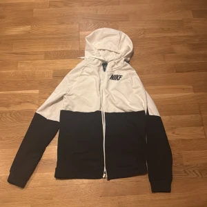 Svart och vit huvtröja från Nike - Säljer en snygg svart och vit huvtröja från Nike med dragkedja. Tröjan har långa ärmar och en luva, perfekt för en sportig look. Tillverkad i Dri-FIT-material för komfort och funktionalitet.