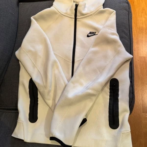 Ljus beige Nike tech kofta - Säljer en stilren vit kofta från Nike med svart dragkedja och logga. Oanvänd och väldigt snygg. Pris går att diskuteras. 