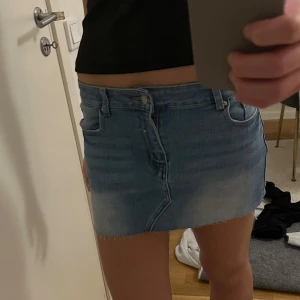 Blå jeanskjol - Säljer en snygg blå jeanskjol med klassisk femficksdesign och knappgylf. Kjolen har en kort längd och en lätt tvättad look som ger en avslappnad stil. Perfekt för varma dagar! Köpt i Spanien. Den är stretchig