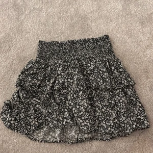 Svart blommig kjol med volang från designers remix - Snygg svart kjol från designers remix med vita blommor och volangdetaljer. Den har en elastisk midja för bekväm passform. Perfekt för att ge en romantisk touch till din outfit.