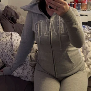 Grå hoodie från GAP - Säljer en mysig grå hoodie från GAP med dragkedja och justerbar huva. Tröjan har långa ärmar och en klassisk logga på bröstet. Perfekt för en avslappnad stil.