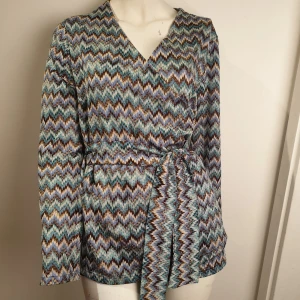 Mönstrad omlottopp från Proud - Snygg omlottopp från Proud, liknar Missioni mönster, med ett färgglatt zigzag-mönster i blått, brunt och beige. Toppen har långa ärmar och ett knytband i midjan för en smickrande passform.  Tillverkad i 100% polyester, perfekt för en trendig look.