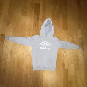 Grå hoodie från Umbro - Säljer en grå hoodie från Umbro med deras klassiska logga tryckt på framsidan. Tröjan har en bekväm huva och långa ärmar, perfekt för kyliga dagar. Storleken är 140
