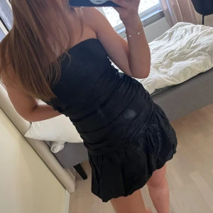 Svart axelbandslös klänning - Snygg svart axelbandslös klänning med en tight passform och volangdetaljer vid fållen. Perfekt för en kväll ute eller en speciell tillställning. Klänningen har en kort längd och ger en elegant look.