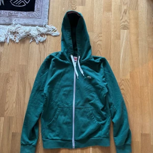 Grön hoodie från Lacoste Live - Säljer en snygg grön hoodie från Lacoste med dragkedja och vit snörning i huvan. Den har en klassisk krokodil-logga på bröstet och ribbade muddar. Perfekt för en avslappnad stil.