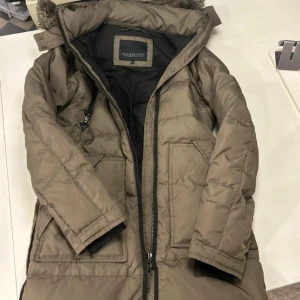 Olivgrön dunjacka från Modström - Säljer en olivgrön dunjacka från Modström med avtagbar huva och fuskpälsdetaljer. Jackan har dragkedja och knappar framtill samt flera praktiska fickor. Perfekt för kalla dagar med sin långa passform och vadderade design. Stl S