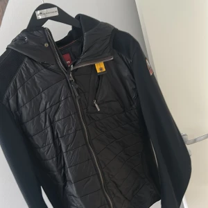 Svart hybridjacka från Parajumpers - Säljer en svart hybridjacka från Parajumpers med quiltad front, huva och dragkedja. Jackan har en gul detalj vid bröstfickan och Parajumpers-logga på ärmen. Perfekt för lager-på-lager under kyliga dagar.