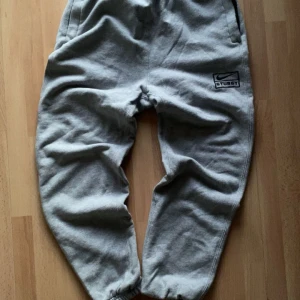 Grå mjukisbyxor från Nike x Stussy - Säljer ett par grå mjukisbyxor från Nike x Stussy. Byxorna har en elastisk midja och muddar vid bensluten för en bekväm passform. De har en broderad logga på sidan och är perfekta för en avslappnad stil.