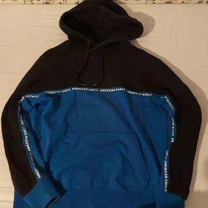 Svart och blå hoodie från American Eagle - Säljer en snygg svart och blå hoodie från American Eagle. Den har en huva och en stor ficka framtill. Detaljer med American Eagle-logga längs ärmarna och bröstet ger en cool touch. Perfekt för en avslappnad stil.