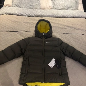 Mörkgrön dunjacka från Sail Racing - Säljer en mörkgrön dunjacka från Sail Racing med gul insida. Jackan har en huva och dragkedja framtill. Perfekt för kalla dagar med sin pufferdesign som håller värmen. Märkesetikett synlig på bröstet.