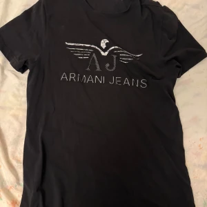 Svart t-shirt från Armani Jeans - Snygg svart t-shirt från Armani Jeans med deras ikoniska logga i silver på bröstet. Perfekt för en stilren look. T-shirten har korta ärmar och en klassisk passform.
