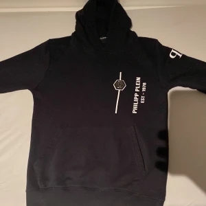 Svart hoodie från Philipp Plein - Snygg svart hoodie från Philipp Plein med tryck på framsidan och en stor ficka. Tillverkad i Italien av bomull och elastan för en bekväm passform. Perfekt för en stilren look.
