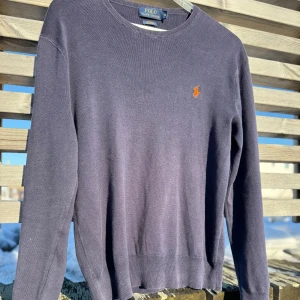 Mörkblå tröja från Ralph Lauren - Säljer en stilren mörkblå tröja från Ralph Lauren med det klassiska broderade logotypen i orange på bröstet. Tröjan har långa ärmar och en rund halsringning. Perfekt för en avslappnad men ändå elegant look.