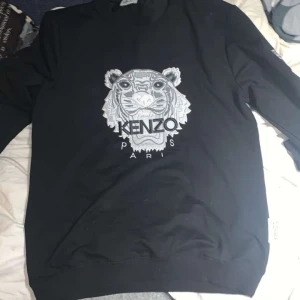 Svart sweatshirt från Kenzo med tigertryck - Snygg svart sweatshirt från Kenzo med ett stort tigertryck i grått och vitt på framsidan. Tröjan har långa ärmar och en klassisk rund halsringning. Perfekt för en stilren och trendig look.
