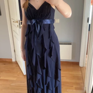 Mörkblå balklänning - Vacker mörkblå långklänning med v-ringad design och volanger. Klänningen har en vacker satinrosett i midjan som ger en fin silhuett. Perfekt för balen eller bröllopsgäst. Passar s men är lite stretchig så kan passa m också. Öppen för förslag
