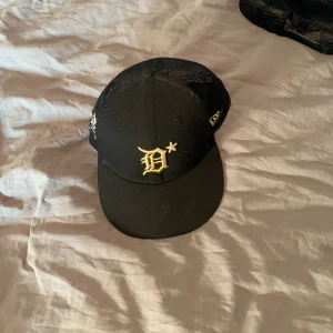 Svart fitted cap - Snygg svart keps med en broderad bokstav i guld på framsidan. Kepsen har ett nätmaterial på baksidan för bättre ventilation. Perfekt för en cool och avslappnad stil.