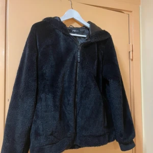 Svart teddyjacka från Fila - Mysig svart teddyjacka från Fila med huva och dragkedja framtill. Perfekt för kyliga dagar när du vill hålla dig varm och bekväm. Jackan har en avslappnad passform och är supermjuk att ha på sig.