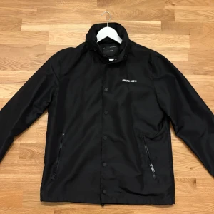 Axel Arigato coach/vind-jacka - Snygg svart jacka från Axel Arigato med tryck 'ARIGATO ©2014' på bröstet och ryggen. Jackan har dragkedja och knappar framtill samt två fickor med dragkedja. Perfekt för en stilren look. Även en luva som kan döljas med dragkedja i kragen