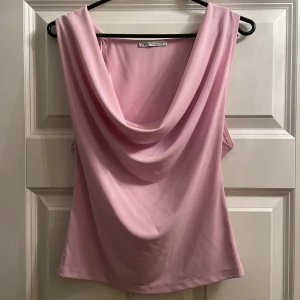 Rosa ärmlös topp från Zara - Säljer en stilren rosa ärmlös topp från Zara med draperad halsringning. Perfekt för en elegant look. Tillverkad i ett mjukt och bekvämt material. I storlek M men passar mig som är 172 cm lång och bär storlek S! Ny utan prislapp aldrig kommit till användning 