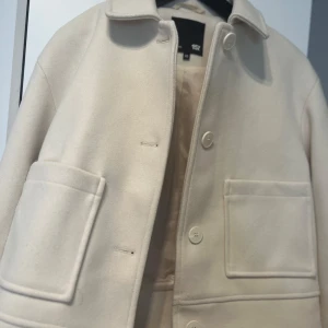 Beige kappa med stora fickor - Snygg beige kappa med stora framfickor och knappar framtill. Jackan har en klassisk krage och långa ärmar med knappdetalj vid ärmsluten. Perfekt för en stilren look. Ny pris 400kr pris går att diskutera 