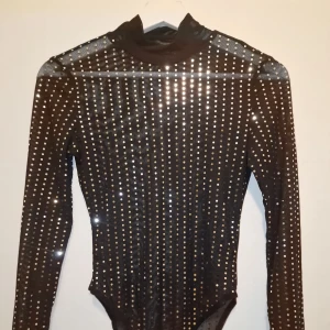 Svart glittrig bodysuit - Snygg svart bodysuit med långa ärmar och glittriga detaljer. Perfekt för en kväll ute eller en speciell tillställning. Garderobsrensning så kika gärna på övriga auktioner.