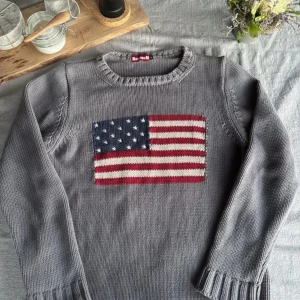 Liknad Ralph Lauren USA tröja - Säljer en grå stickad tröja från Homerun med en stor amerikansk flagga på framsidan. Tröjan har långa ärmar och är tillverkad i 100% bomull. Perfekt för en avslappnad stil.
