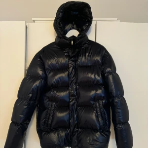 Svart pufferjacka - Säljer en svart pufferjacka med glansig yta och hög krage. Jackan har en dragkedja framtill och långa ärmar. Perfekt för kyliga dagar.