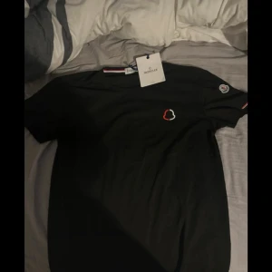 Svart t-shirt från Moncler - Snygg svart t-shirt från Moncler med korta ärmar. Den har en liten broderad logga i rött och vitt på bröstet och en patch på ärmen. Perfekt för en stilren look.