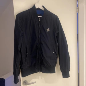 Mörkblå bomberjacka från J.Lindeberg - Snygg mörkblå bomberjacka från J.Lindeberg med dragkedja och ribbade muddar. Jackan har en stilren design med en liten logotyp på bröstet. Perfekt för en cool och avslappnad look.