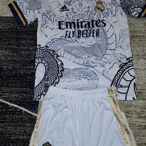 Vit Real Madrid fotbollströja med drakmönster. Cristiano Ronaldo nummer 7 - Säljer en vit Real Madrid fotbollströja från Adidas med ett coolt drakmönster. Tröjan har korta ärmar och klubbens emblem på bröstet. Den har även sponsorlogga och spelarnamn på ryggen. Perfekt för fans som vill sticka ut på planen!