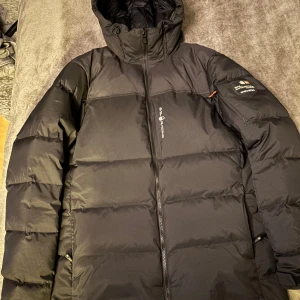 Sail racing Jacka  - Säljer en svart dunjacka från Sail Racing med huva och dragkedja. Jackan har en broderad logga på ärmen och är perfekt för kalla dagar. Den är långärmad och designad för att hålla dig varm i extrema förhållanden. Använd 3-4 gånger inte reportage eller skador 