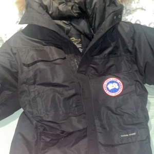 Svart dunjacka från Canada Goose - Säljer en svart dunjacka från Canada Goose med pälsdetalj på huvan. Jackan har en klassisk design med dragkedja och knappar framtill samt en ikonisk logotyp på bröstet. Perfekt för kalla vinterdagar.
