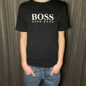 Svart t-shirt från Hugo Boss - Snygg svart t-shirt från Hugo Boss med stort vitt tryck på bröstet. Perfekt för en stilren look. Kortärmad och tillverkad i mjukt material för bekväm passform.