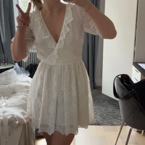 Studentklänning - Charmig vit omlottklänning med v-ringning och spetsdetaljer. Klänningen har trekvartsärmar och ett fint broderat mönster. Perfekt för studenten. 