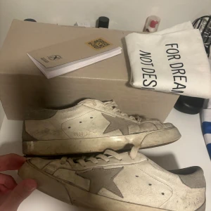 Golden goose - Säljer ett par vita sneakers med en grå stjärndetalj på sidan. Skorna har snörning och en klassisk design. Perfekta för en avslappnad stil.