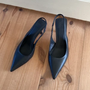 Svarta pumps med klack - Eleganta svarta pumps med spetsig tå och smal klack. Perfekta för en stilren look. Tillverkade i ett slätt material med en enkel remdesign. Lite slitna i klacken men finns nya tillhörande som skickas med!