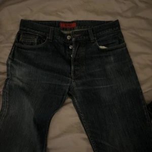 Mörkblå jeans från Hugo Boss - Snygga mörkblå jeans från Hugo Boss med klassisk femficksdesign och knappgylf. Perfekta för en stilren look. Märkeslappen i rött ger en extra touch.