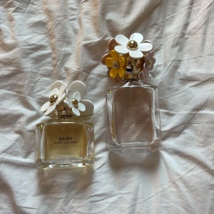 Daisy parfymer från Marc Jacobs - Två eleganta Daisy parfymer från Marc Jacobs. Den ena flaskan har en klar vätska och den andra en ljusgul ton. Båda flaskorna har vackra blomformade lock i vitt, gult och beige med gulddetaljer. Perfekt för den som älskar blommiga och fräscha dofter.