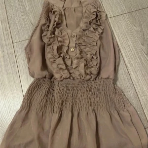 Beige topp volangdetaljer - Säljer en söt beige ärmlös topp med volangdetaljer framtill och knappar. Toppen har en smockad midja som ger en fin passform. Perfekt för varma dagar!