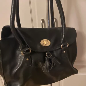 Svart axelväska från Tommy Hilfiger - Snygg svart axelväska från Tommy Hilfiger i skinn. Väskan har en stilren design med guldfärgade detaljer och tofsar framtill. Perfekt för både vardag och fest! 🖤