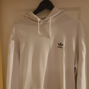 Vit hoodie från Adidas - Säljer en snygg vit hoodie från Adidas i storlek L. Den är i bra skick och har en klassisk design med Adidas-loggan på bröstet. Perfekt för både vardag och träning. Den har en stor ficka fram och en bekväm huva med dragsko. Primegreen-material för en hållbar touch.