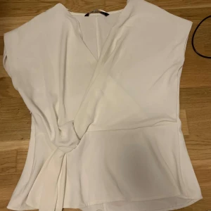 Vit omlottopp - Säljer en stilren vit omlottopp i mjukt material. Toppen har en elegant omlottdesign och är perfekt för både vardag och fest. Den är ärmlös och har en lös passform som ger en bekväm känsla. Passar bra till både jeans och kjol!